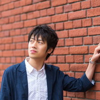 レンガの壁に片肩を寄せて寄りかかるカジュアルな青年の写真