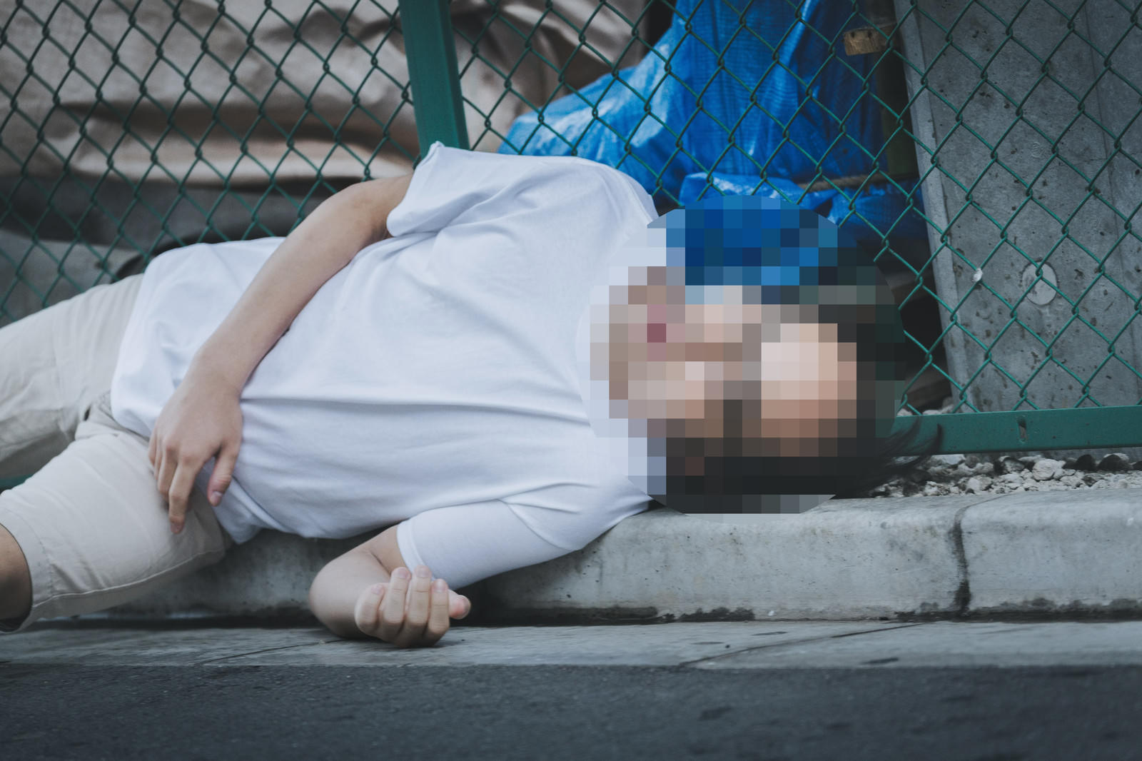 白いシャツの男性が緑のフェンス横のコンクリート路上に倒れ込み、胸に腕を当てている［モデル：モデルリリース］