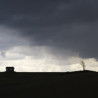 暗い雲と光が差す荒野に建つ一軒家と枯れ木のシルエットの写真