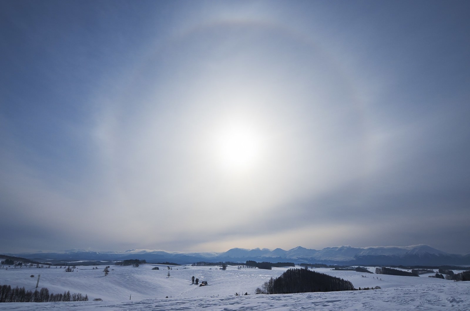 雪に覆われた平原と山並みの上空に太陽を囲む白い光の輪のハロ現象が見える