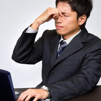 ノートPCの前で鼻の付け根を押さえる目が疲れたビジネスマンの写真