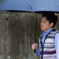 雨の日にコンクリート壁の前で傘を差して恋人を待つ男性の写真