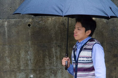 雨の日にコンクリート壁の前で傘を差して恋人を待つ男性
