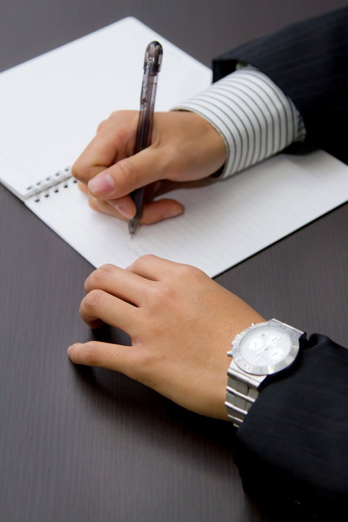 灰色の机でペンを持ちノートにメモを取る両手