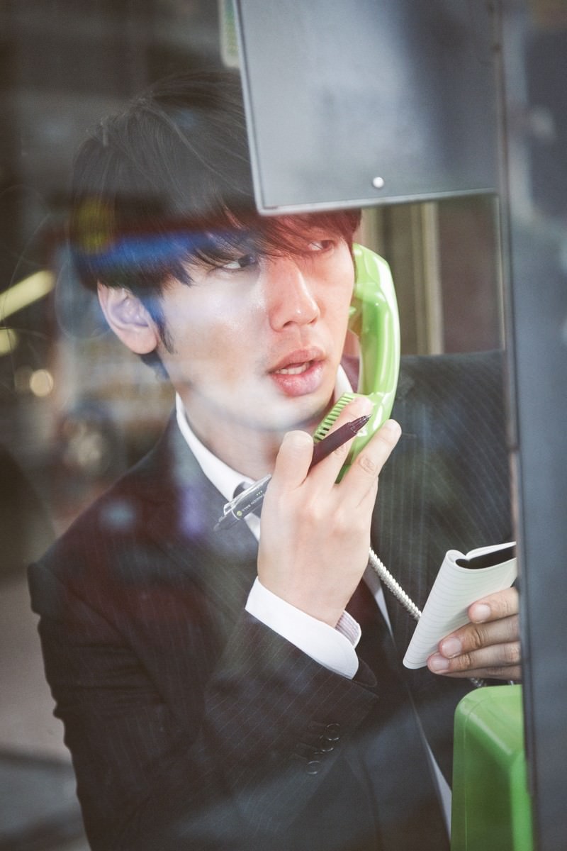 公衆電話ボックスで小銭がないことに気づき下を向く男性ジャーナリスト［モデル：大川竜弥］