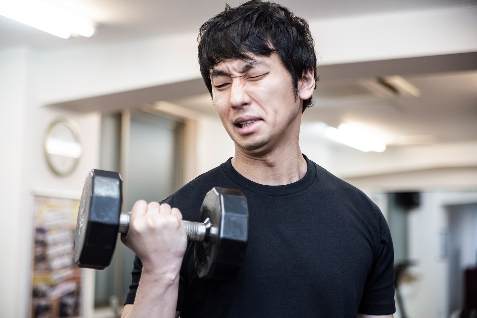 ダンベルを握り目を閉じた男性がジムでトレーニングをしている。疲労した表情で苦しい状態を表現［モデル：大川竜弥］