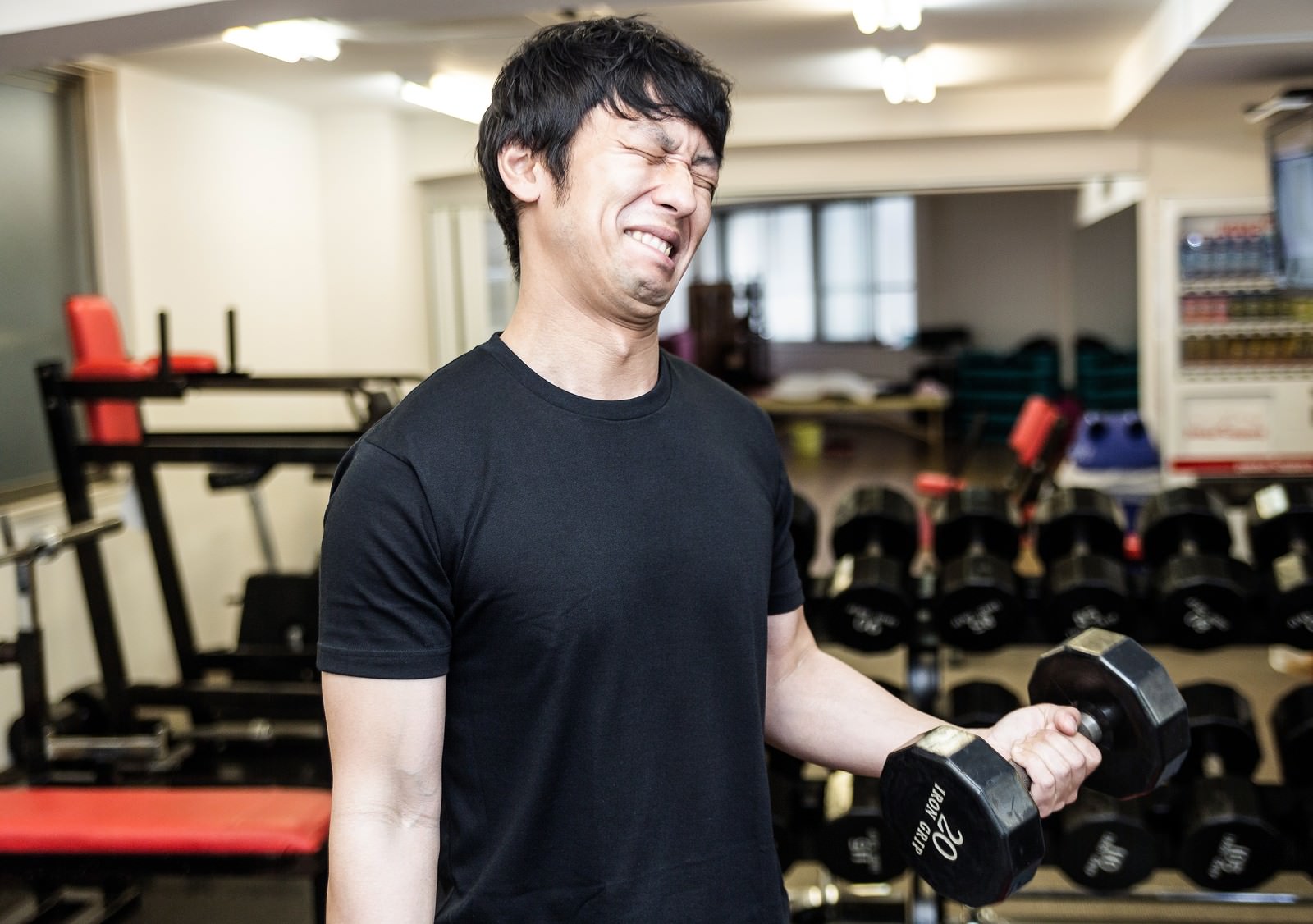 ダンベルを握りしめて歯を食いしばる男性がジムで限界のトレーニングをしている［モデル：大川竜弥］