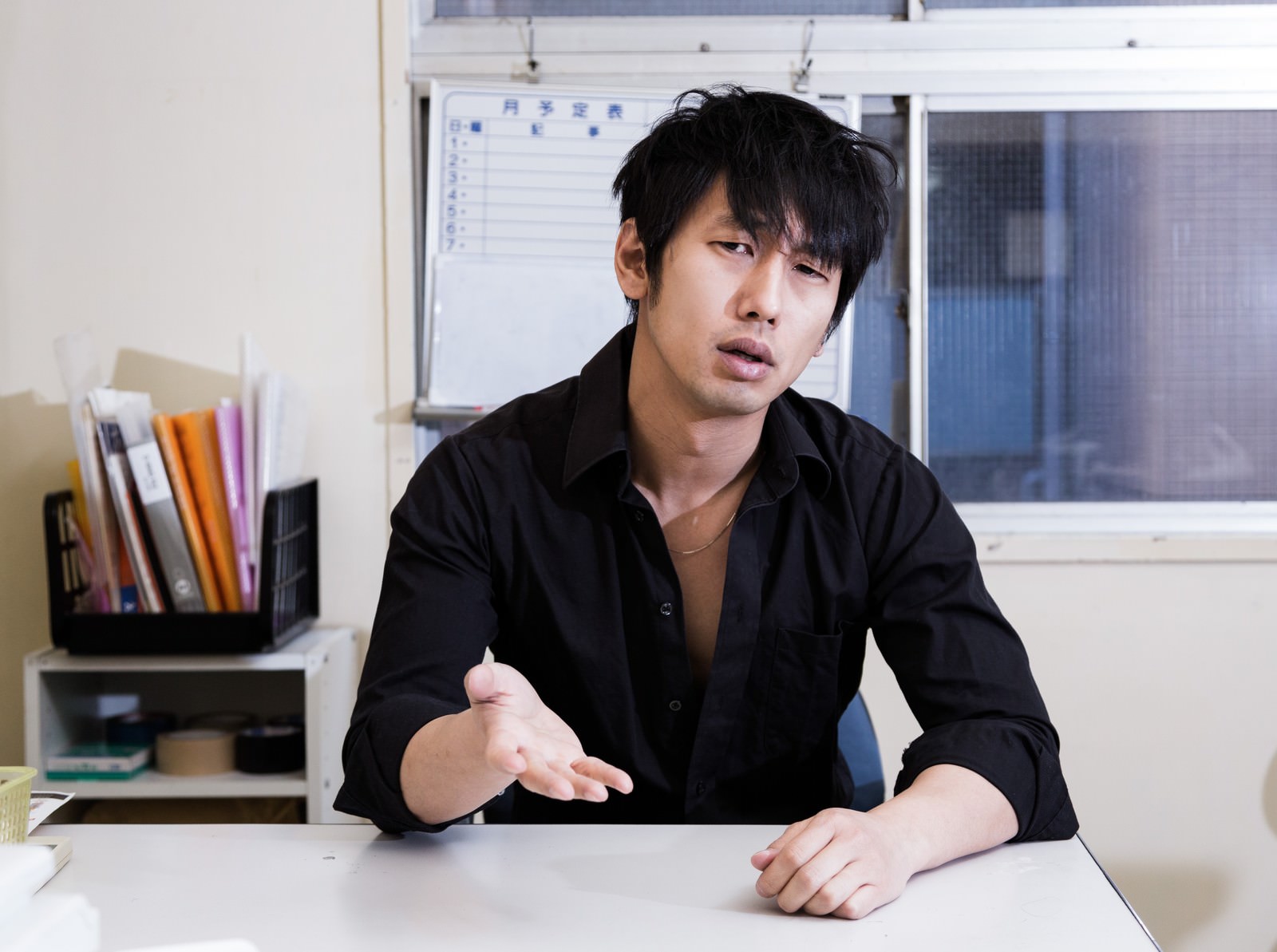 オフィスの机で圧迫面接を受ける黒いシャツ姿の若い男性［モデル：大川竜弥］