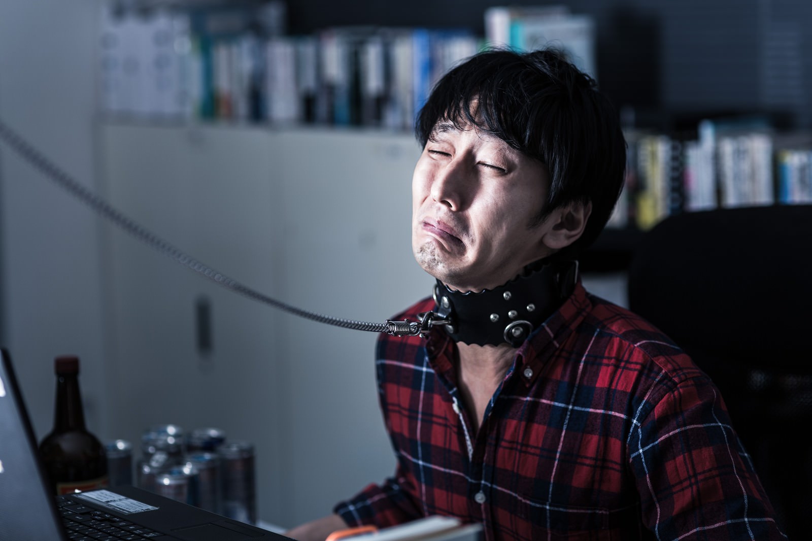 暗い室内で黒い首輪を付けてノートPCの前に座るチェック柄シャツの男性［モデル：大川竜弥］