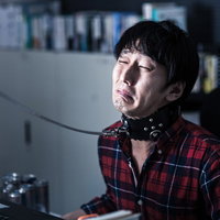 暗い室内で黒い首輪を付けてノートPCの前に座るチェック柄シャツの男性の写真