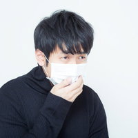白いマスクを右手で押さえる黒い襟付きニットの男性の写真