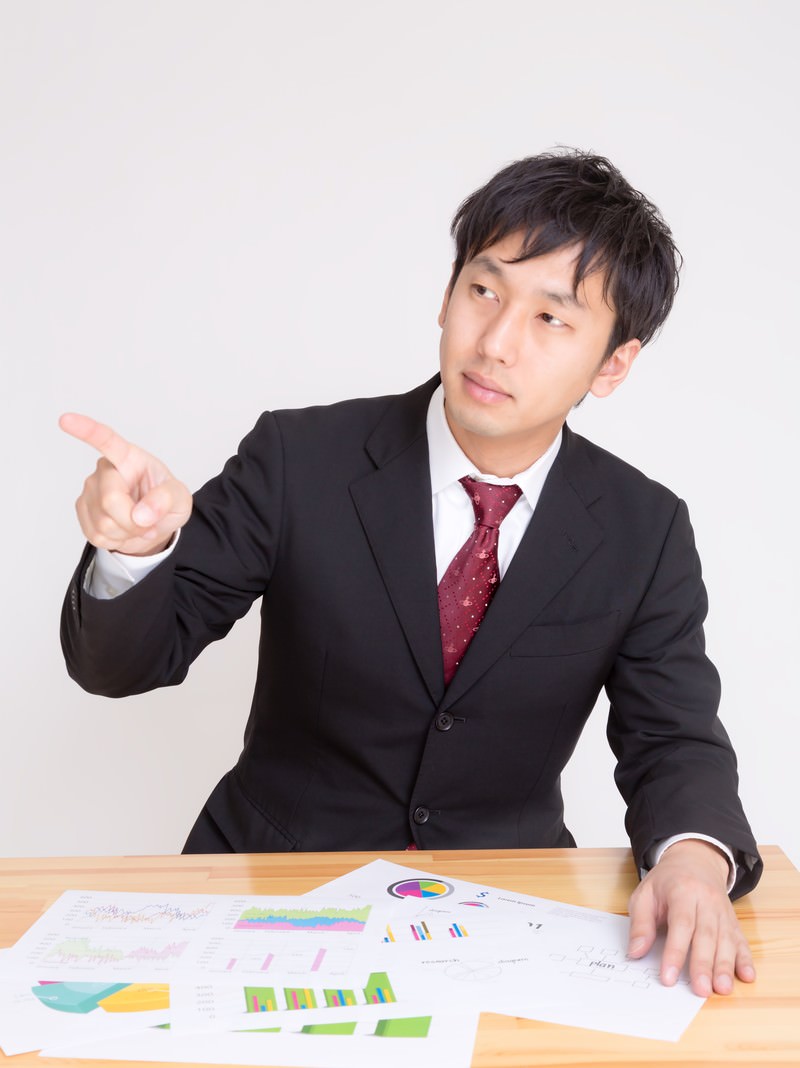 スーツ姿の男性がテーブルの上のグラフを指して説明している［モデル：大川竜弥］