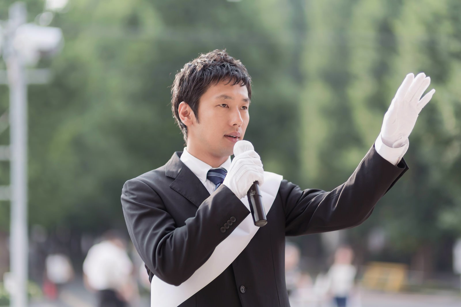白い手袋でマイクを持ち街頭演説する男性［モデル：大川竜弥］