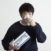 紺色のセーターでティッシュを鼻に当てもう片手にティッシュ箱を持つ黒髪の人物の写真