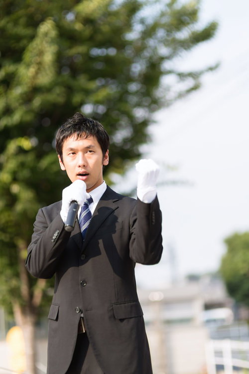 屋外でマイクと紙を持って演説するスーツの男性