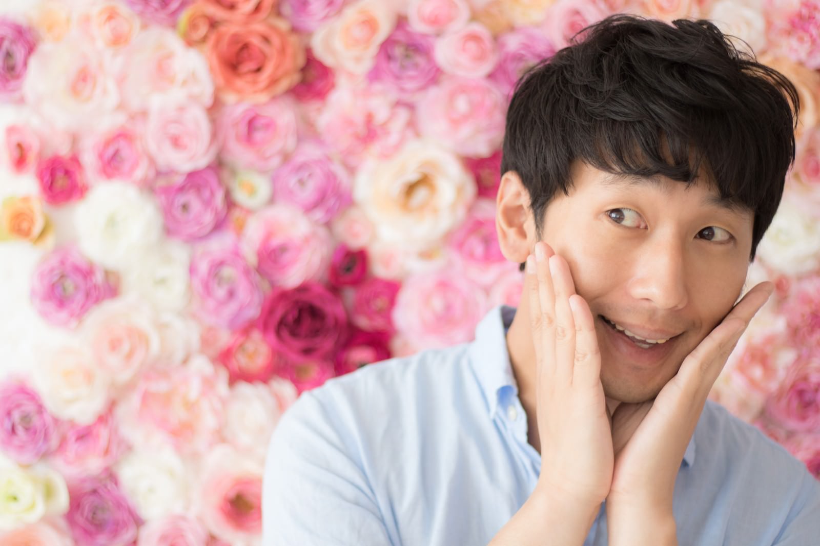 ピンク色のバラが咲く花壁を背景に両手を顔に添えて笑顔を見せる男性［モデル：大川竜弥］