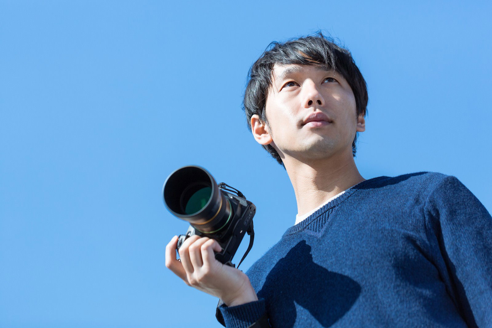 青空を背景に、望遠レンズ付きの一眼レフカメラを構えるカメラ男子［モデル：大川竜弥］