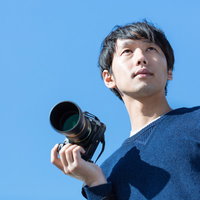 お気に入りの一眼レフカメラを持って青空の下に立つカメラ男子の写真