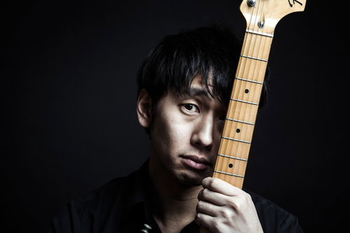 暗い室内でメイプル指板のベースのネックを持ち正面を見る黒髪男性