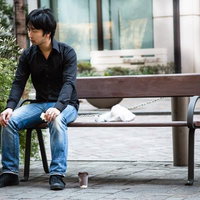 建物前のベンチに座り足元にテイクアウトカップを置く黒シャツの黒髪の人物の写真
