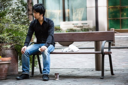 建物前のベンチに座り足元にテイクアウトカップを置く黒シャツの黒髪の人物