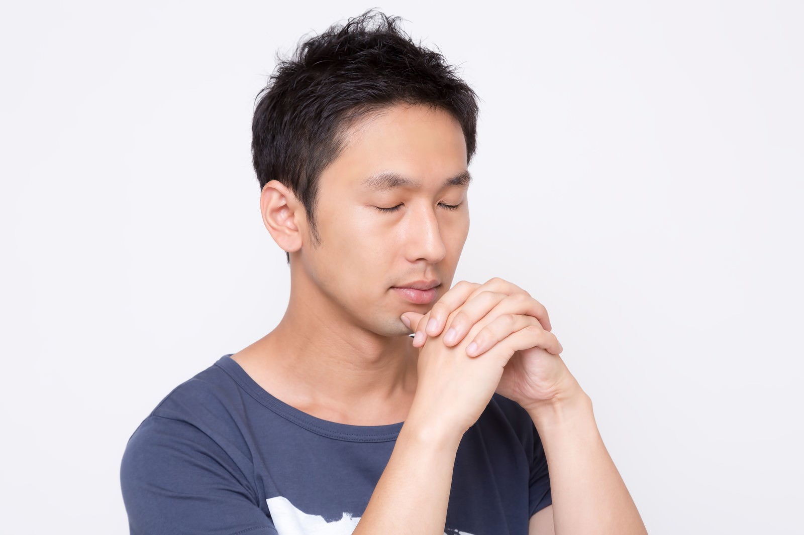 紺色のTシャツを着た男性が両手を胸の前で合わせて目を閉じ、祈るような姿勢をしている［モデル：大川竜弥］