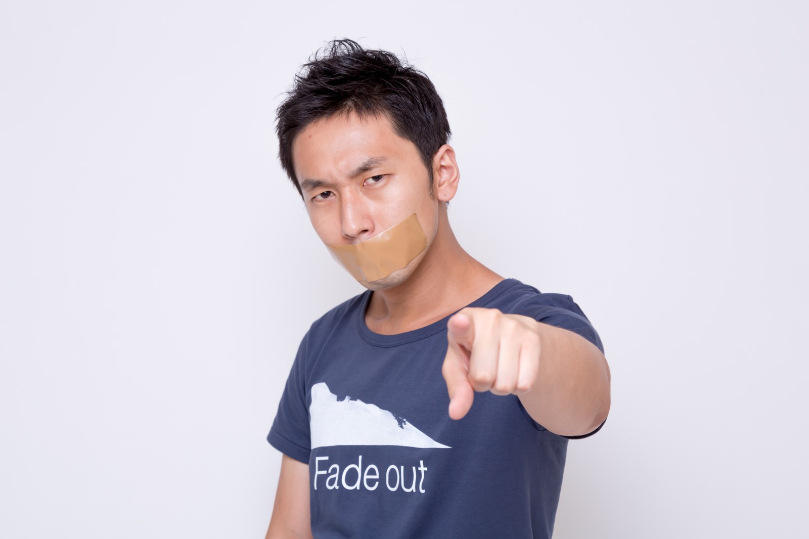 口にガムテープを貼られて喋れない男性がカメラに向かって人差し指を突き出している［モデル：大川竜弥］