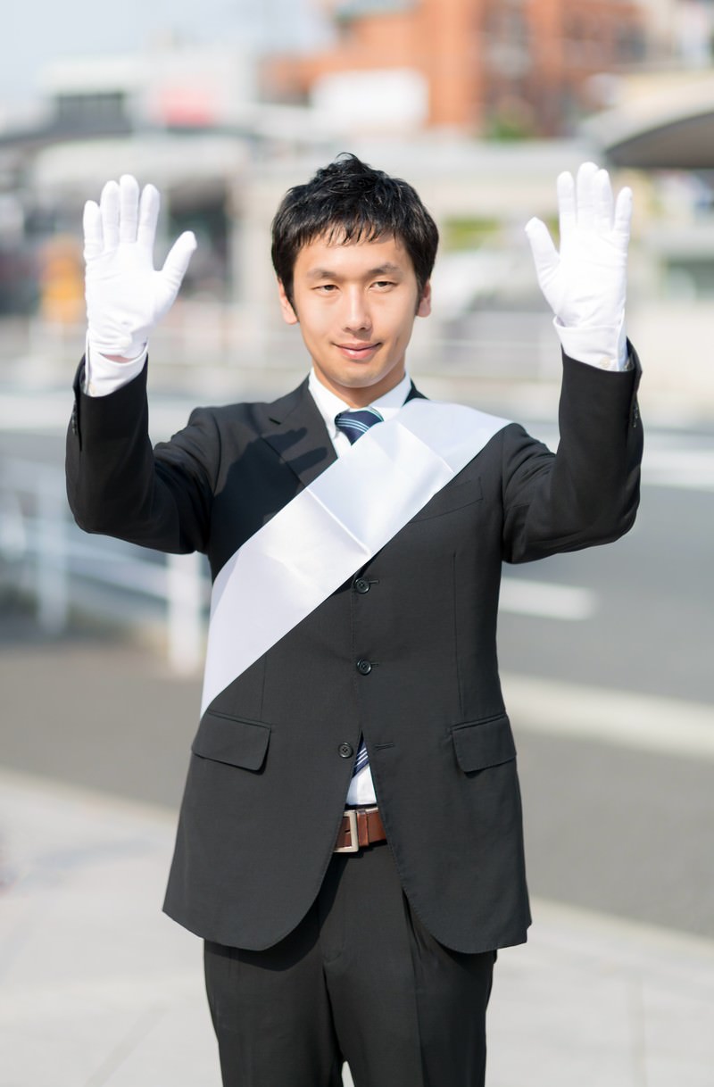 たすきをかけて両手を挙げる党公認候補［モデル：大川竜弥］