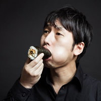 暗い背景で右手に巻き寿司を持って口に運ぶ黒い服の男性の写真