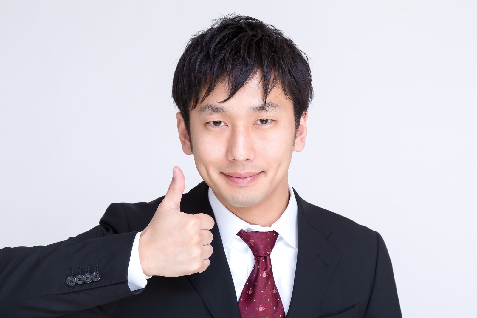 黒いスーツに白シャツ、えんじ色ネクタイを着た男性がサムズアップを見せながら笑顔で微笑む［モデル：大川竜弥］