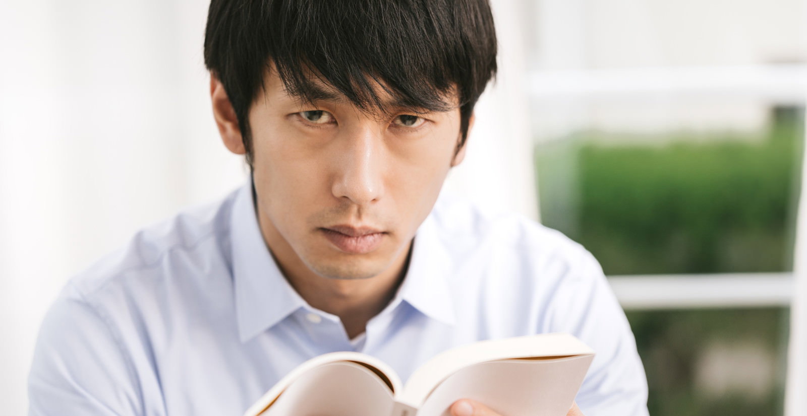 白いシャツの男性が机で本を両手で持ち読んでいる［モデル：大川竜弥］