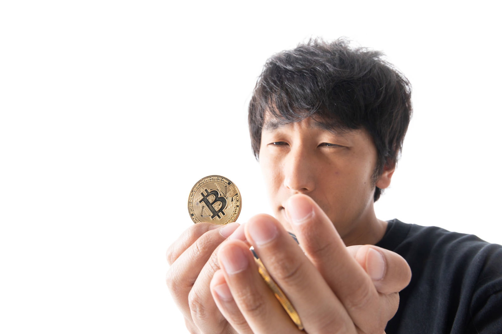 金色のビットコインコインを指先で持って見つめる黒いシャツの男性［モデル：大川竜弥］