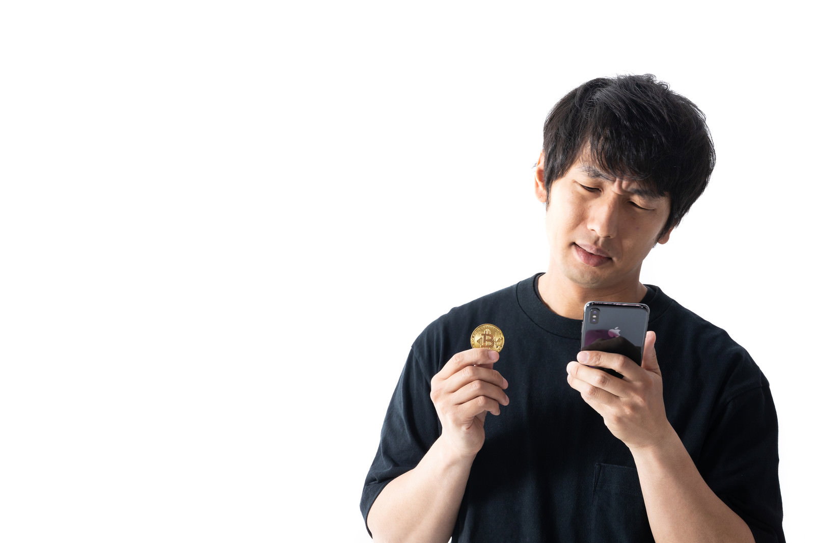 スマートフォンとビットコインを手に持ち、画面を見下ろして困惑する黒い服の男性［モデル：大川竜弥］