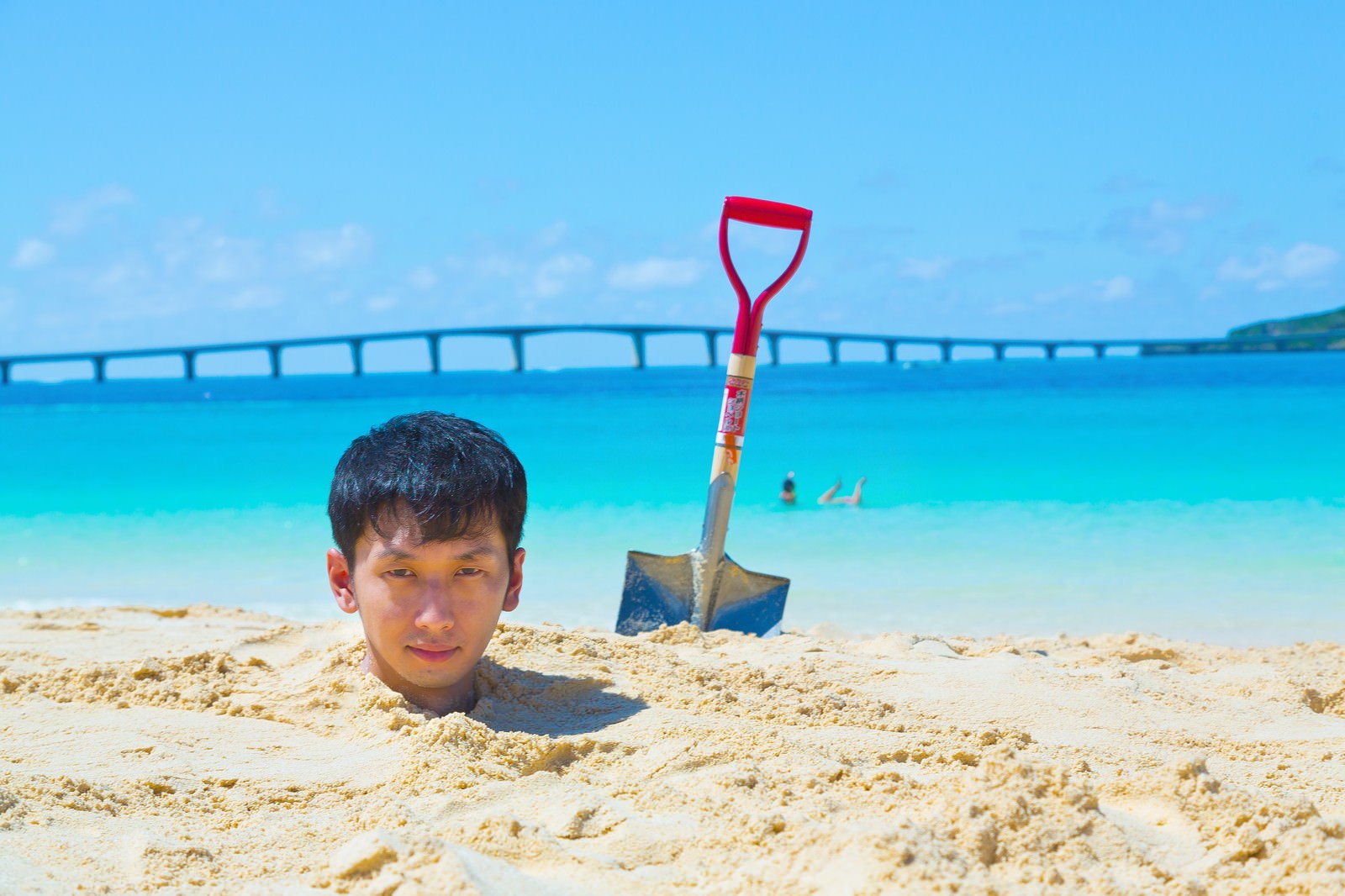 宮古島の砂浜で砂に埋められ顔を出す少年と、立てられた赤いスコップ、背景の海と橋［モデル：大川竜弥］