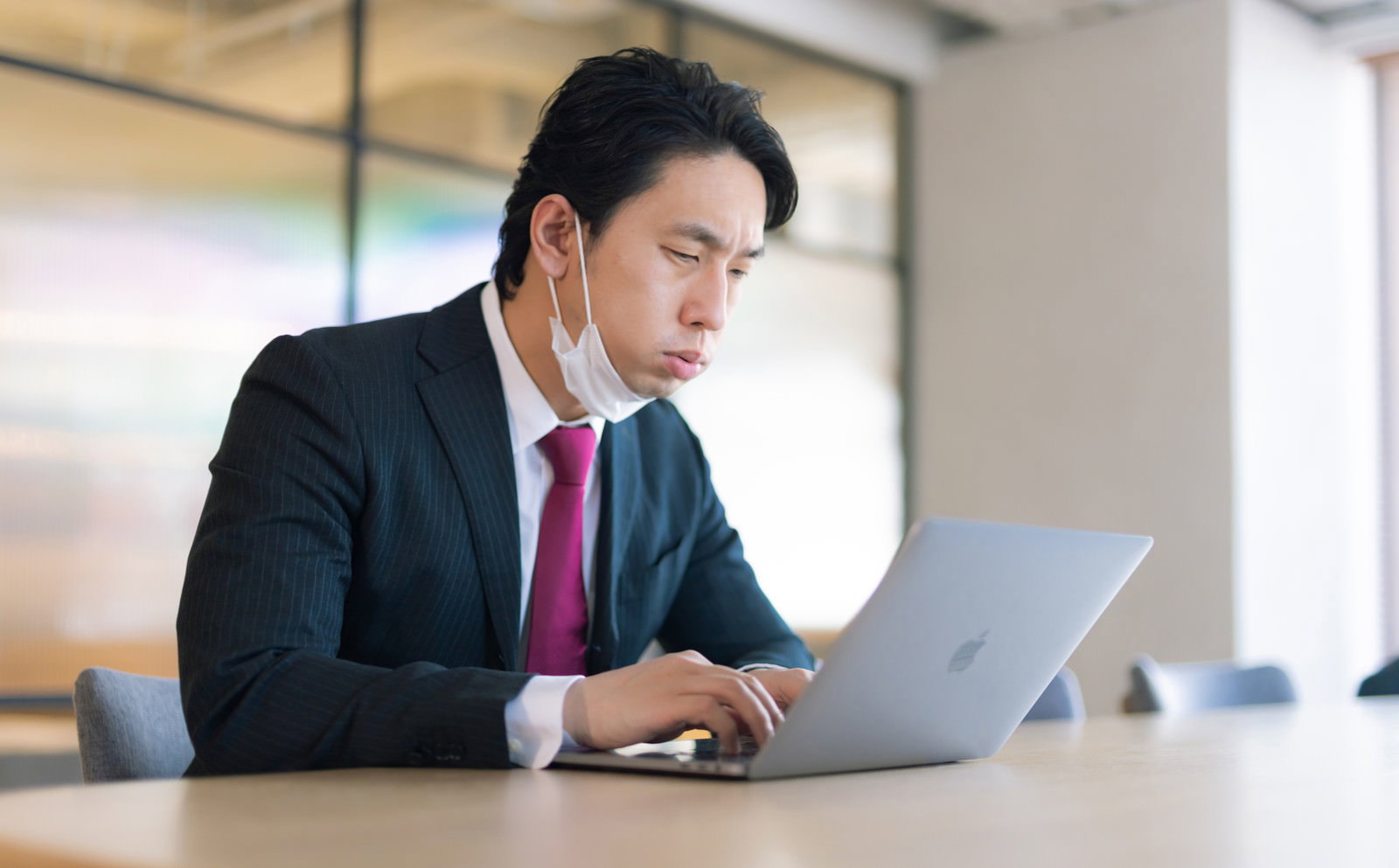 オフィスでノートパソコンに向かい不満そうな表情で作業するスーツ姿の会社員男性［モデル：大川竜弥］