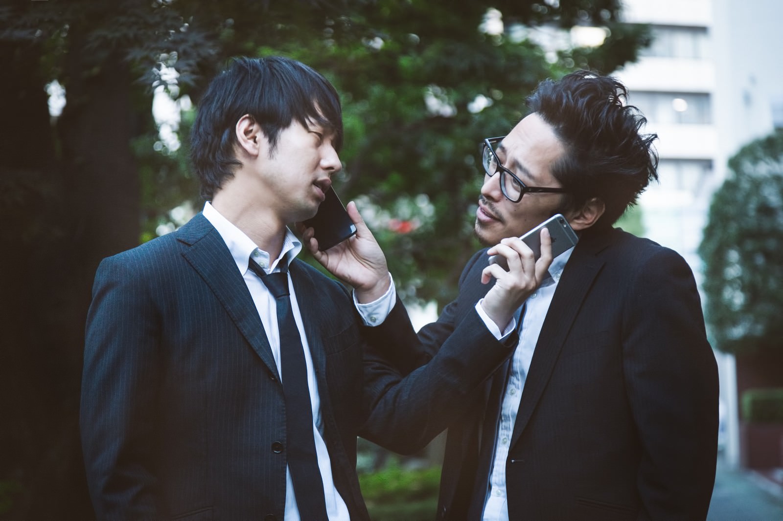スーツ姿の2人の男性が屋外で携帯電話とスマートフォンを持ちながら向かい合って話す［モデル：ゆうせい 大川竜弥］