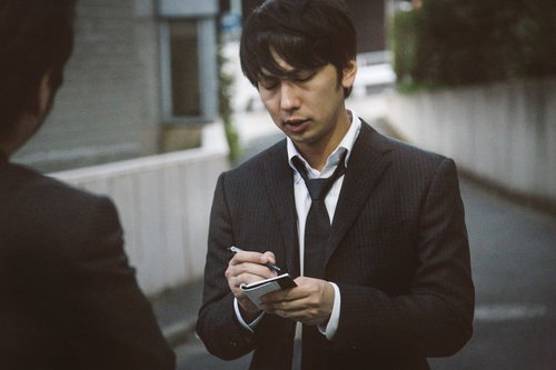 犯行現場付近で聞き込み取材をする若手記者が手帳にメモを取る