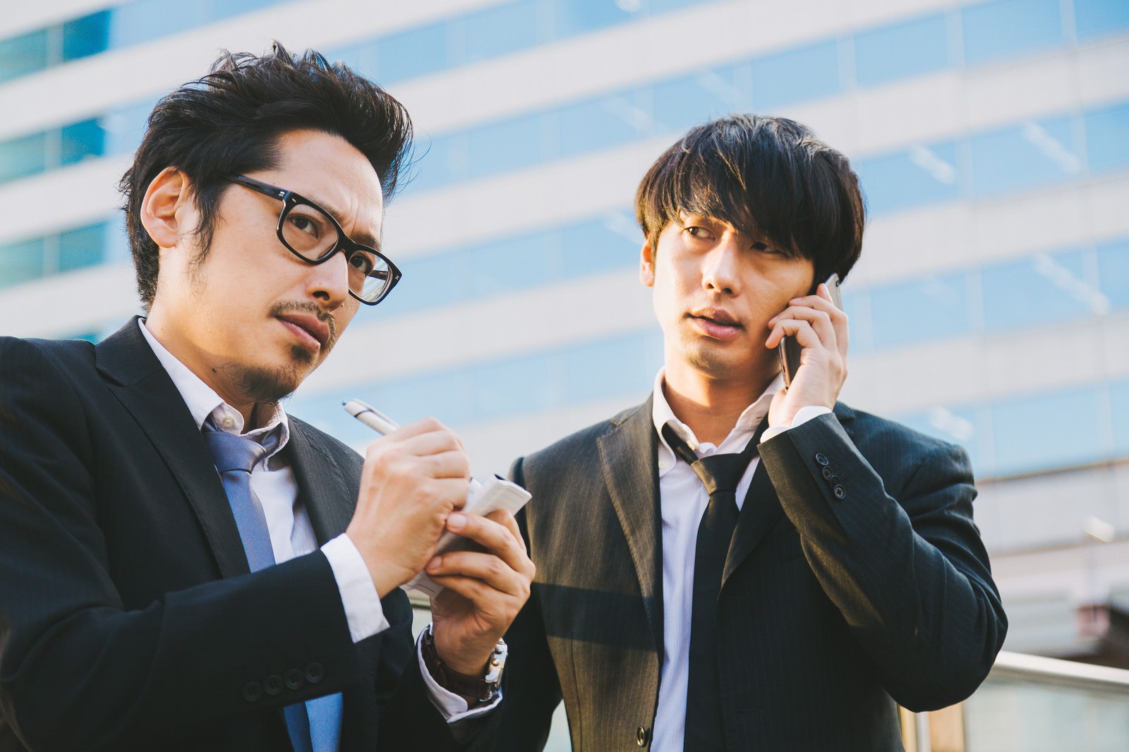 ビルを背景に、ノートとペンを持つ男性と、スマートフォンで通話する男性［モデル：ゆうせい 大川竜弥］