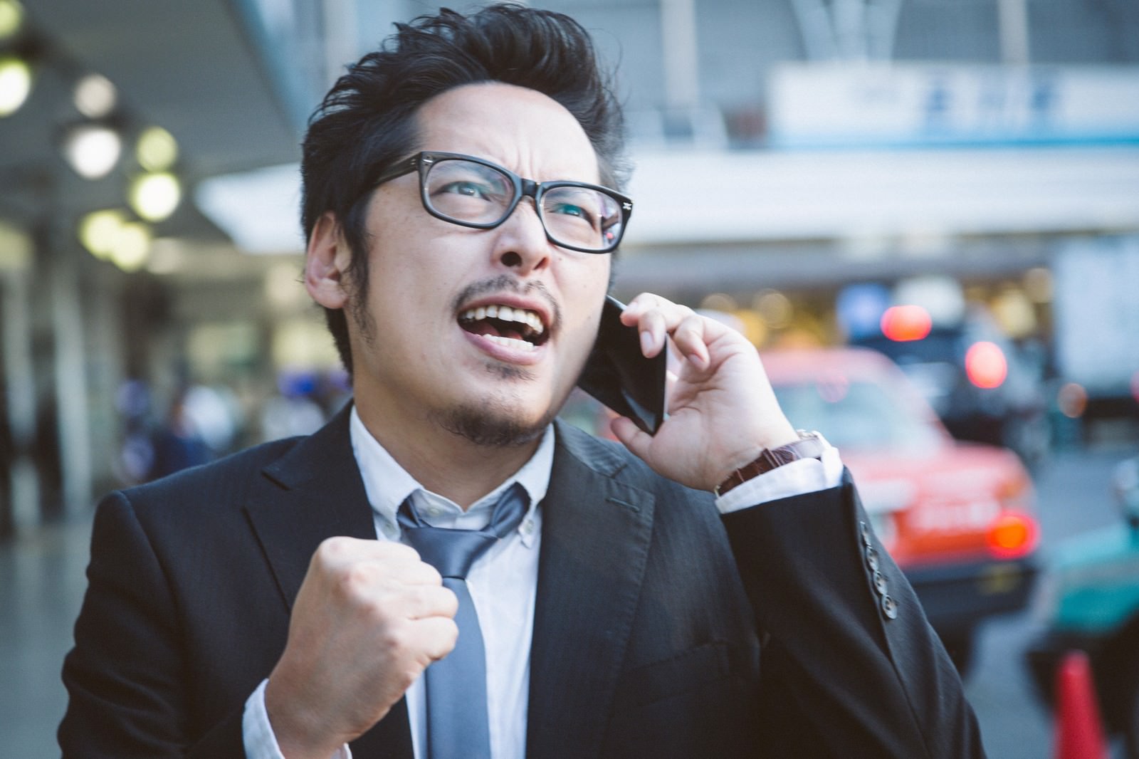 スマートフォンを持ってガッツポーズをする眼鏡をかけたスーツ姿の男性［モデル：ゆうせい］