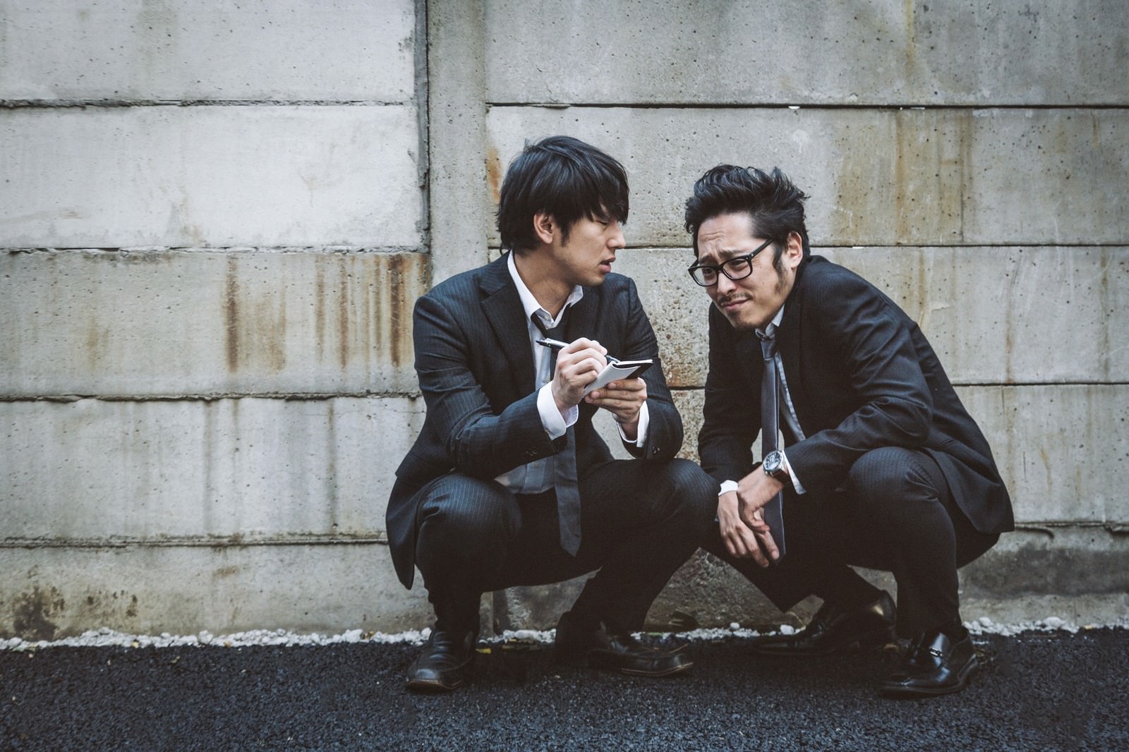 コンクリートの壁の前で蹲って話し合う二人の男性ジャーナリスト［モデル：大川竜弥 ゆうせい］