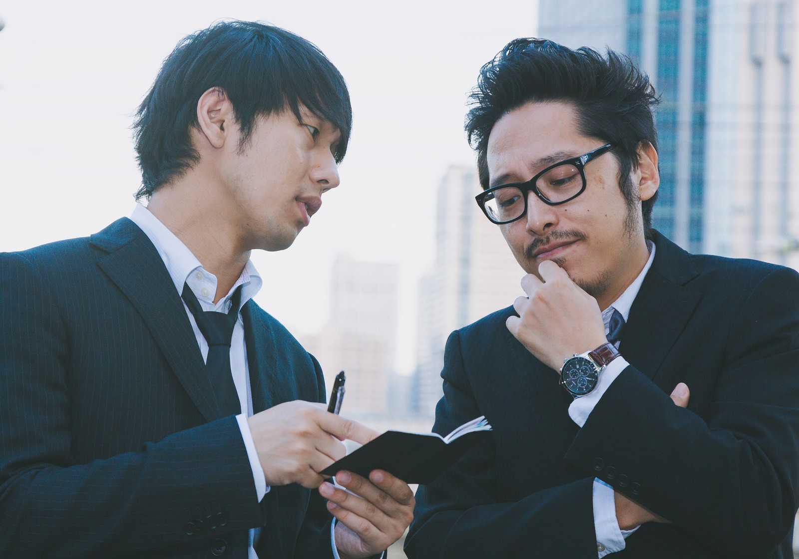 スーツを着た男性二人がビジネスビルを背景に、メモ帳を見ながら取材について打ち合わせしている［モデル：大川竜弥 ゆうせい］