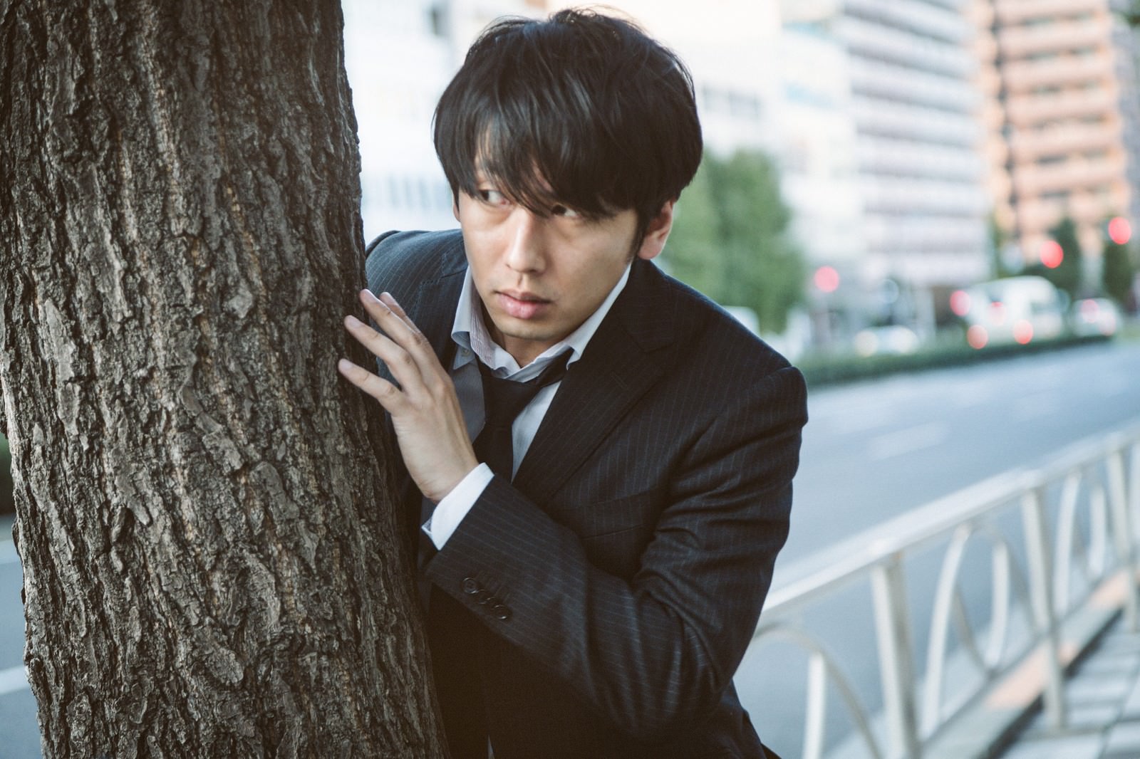 木陰に隠れるスーツ姿の男性がビル群の背景で幹に手を添えている［モデル：大川竜弥］