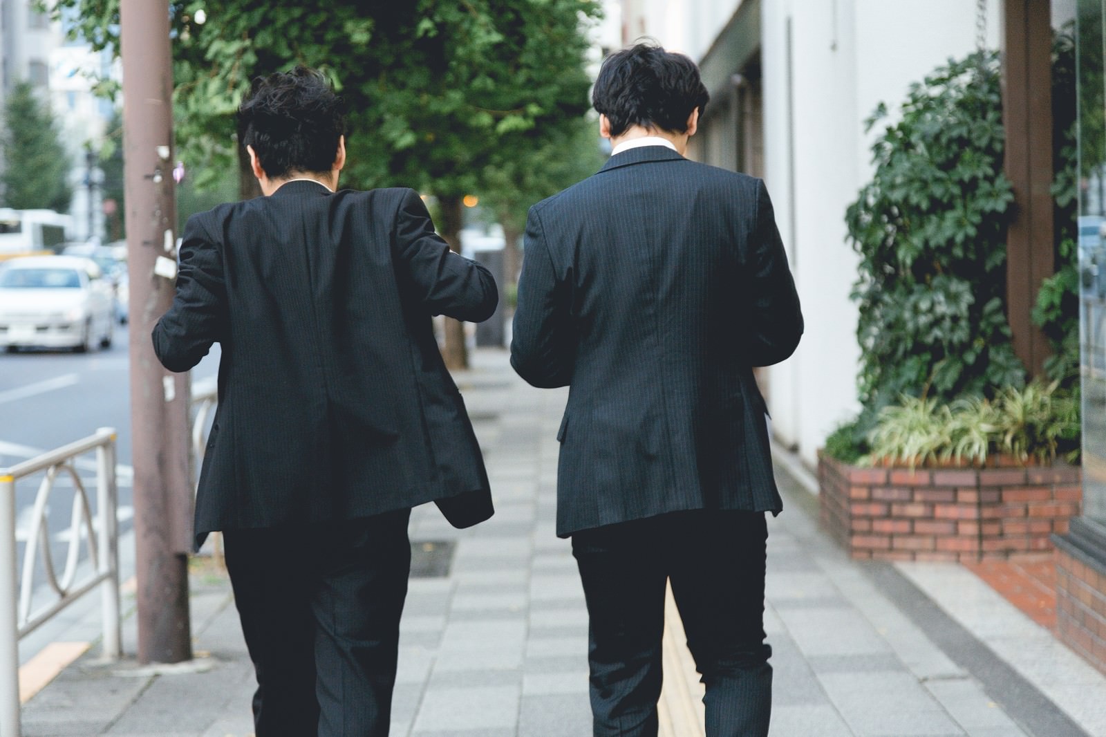 紺色のスーツを着た男性2人が肩を寄せて歩く後ろ姿。背景は緑と建物に囲まれた街路［モデル：ゆうせい 大川竜弥］