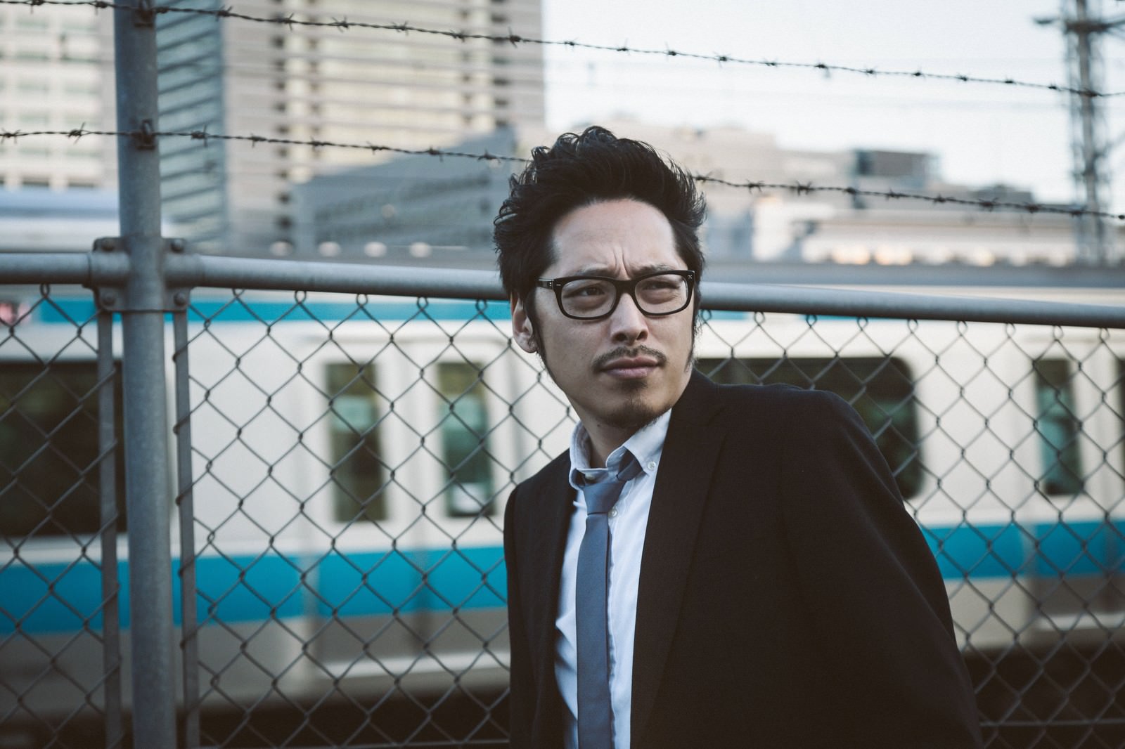 黒いスーツ姿でフェンス越しに張り込み調査をするジャーナリストの男性［モデル：ゆうせい］