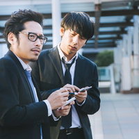 取材中にスマートフォンで時間をつぶす眼鏡をかけた新人記者と同僚の写真