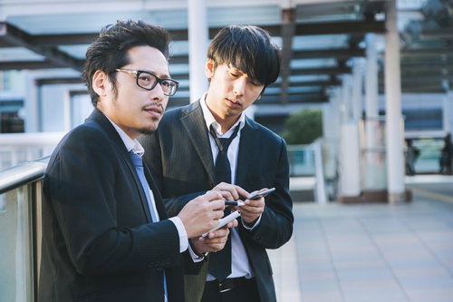 取材中にスマートフォンで時間をつぶす眼鏡をかけた新人記者と同僚