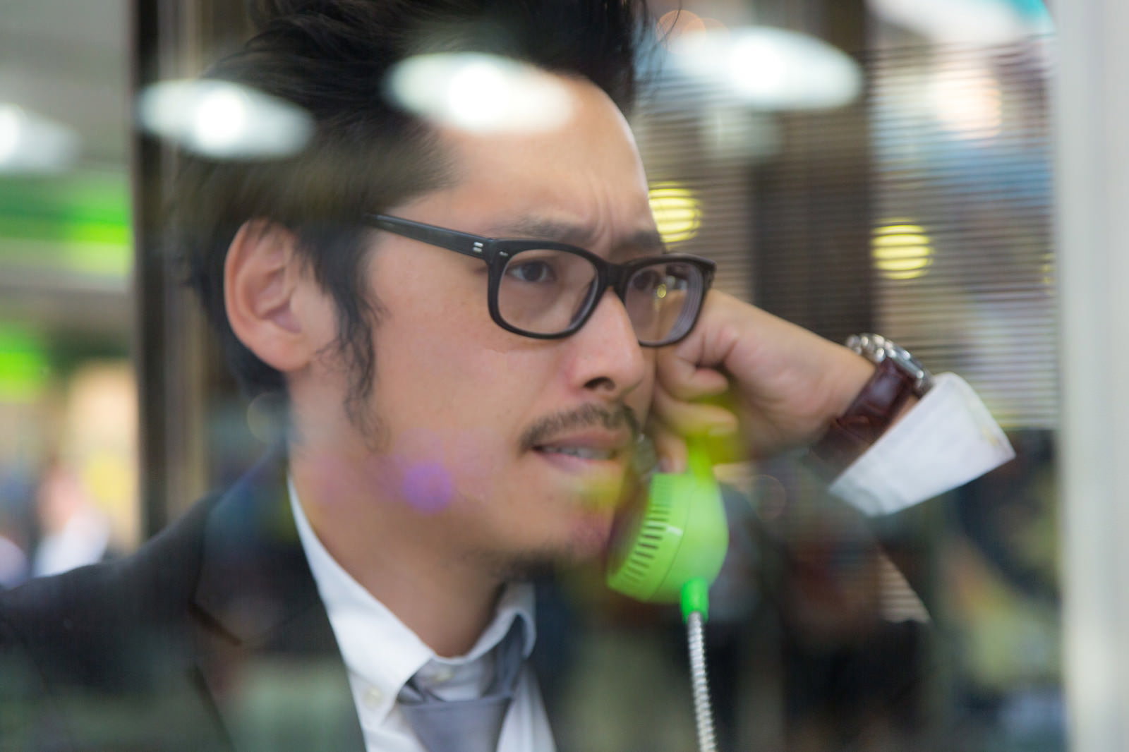 電話ボックスで公衆電話を使って通話する眼鏡をかけたスーツ姿の男性［モデル：ゆうせい］
