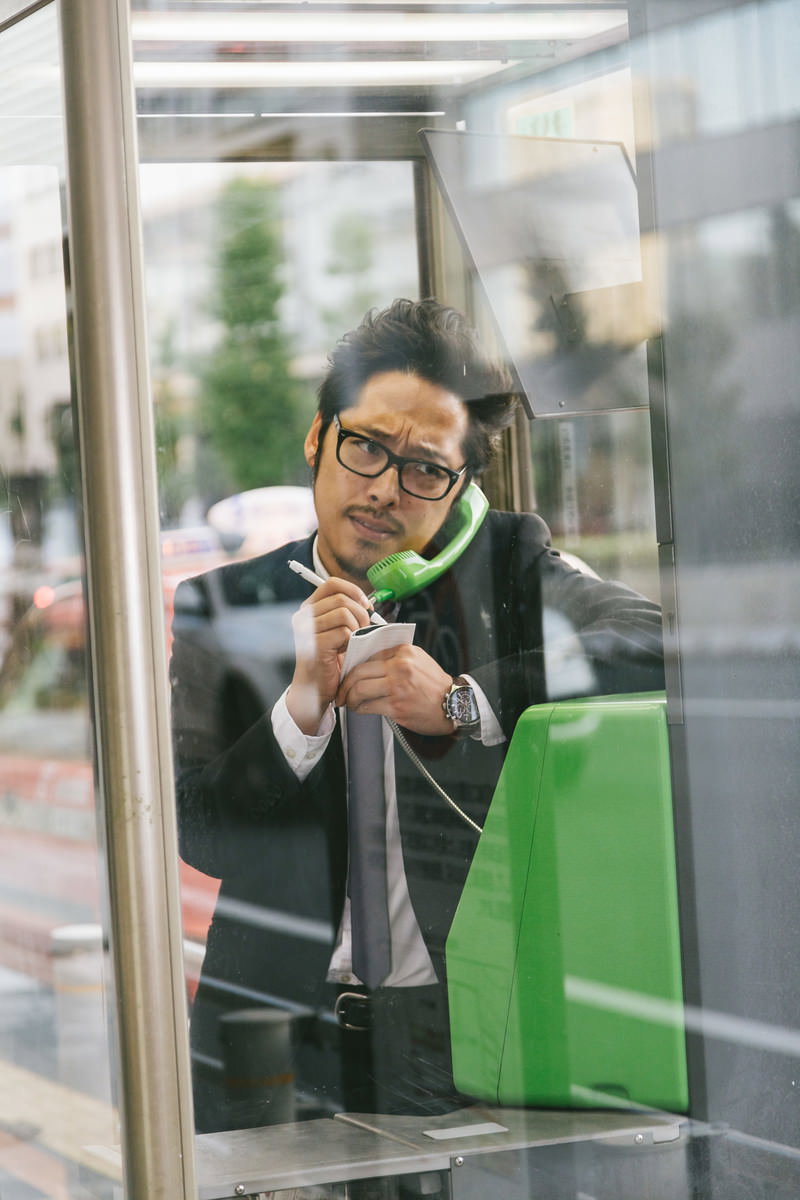 公衆電話ボックス内でメモを取りながら通話するスーツ姿の男性［モデル：ゆうせい］
