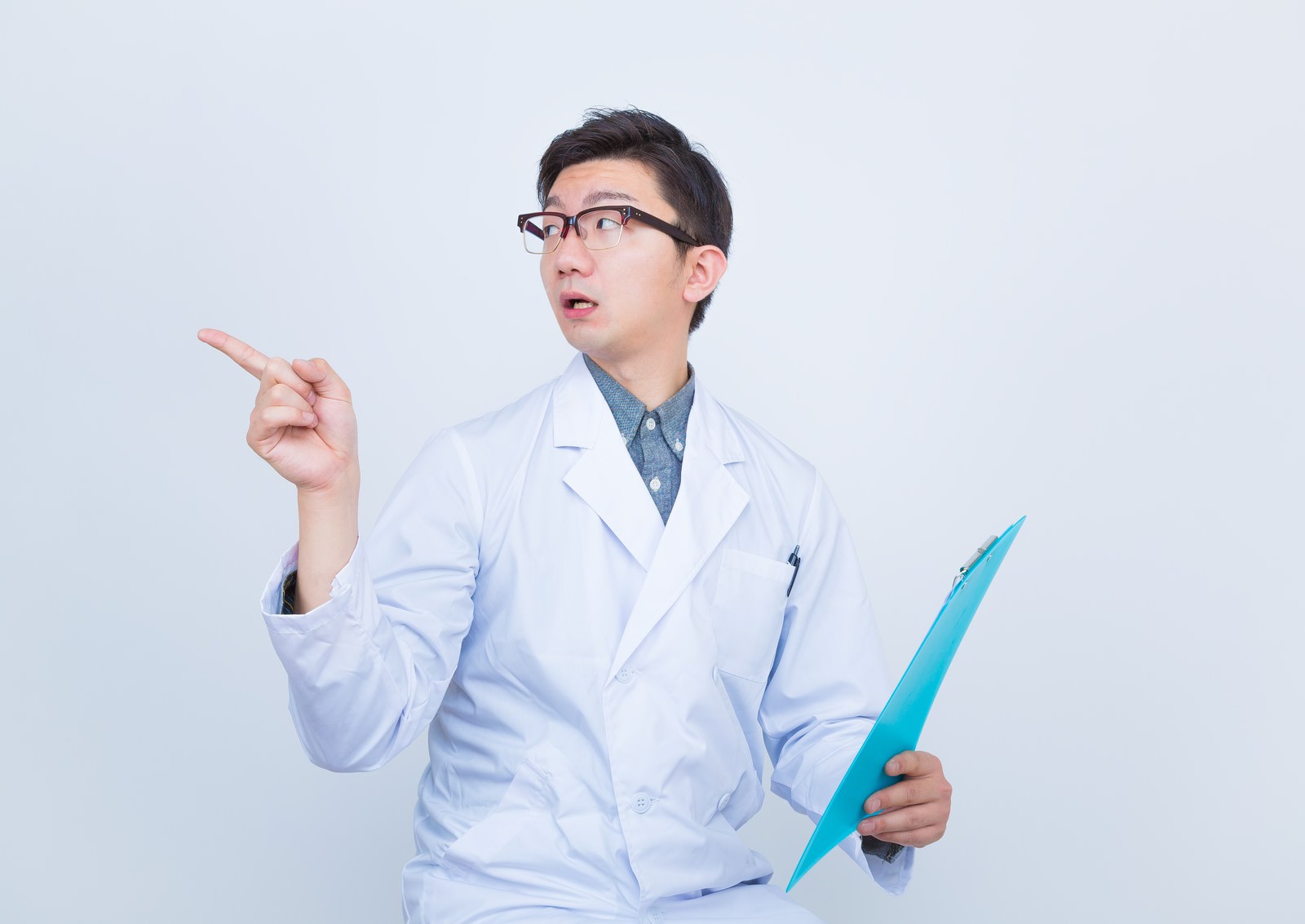 クリップボードを持ち右手で指示ジェスチャーをする白衣姿の眼鏡をかけた男性医師［モデル：OZPA］