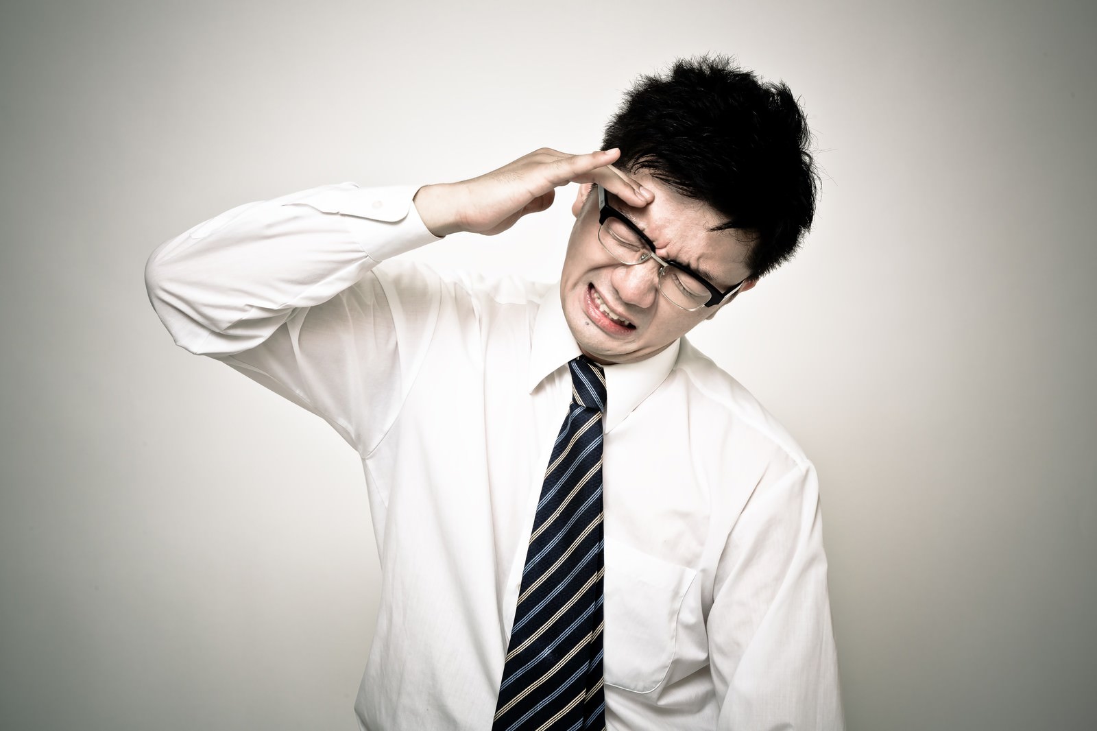 眼鏡の男性がワイシャツとネクタイ姿で右手を額に当て歯を見せた表情をしている［モデル：OZPA］